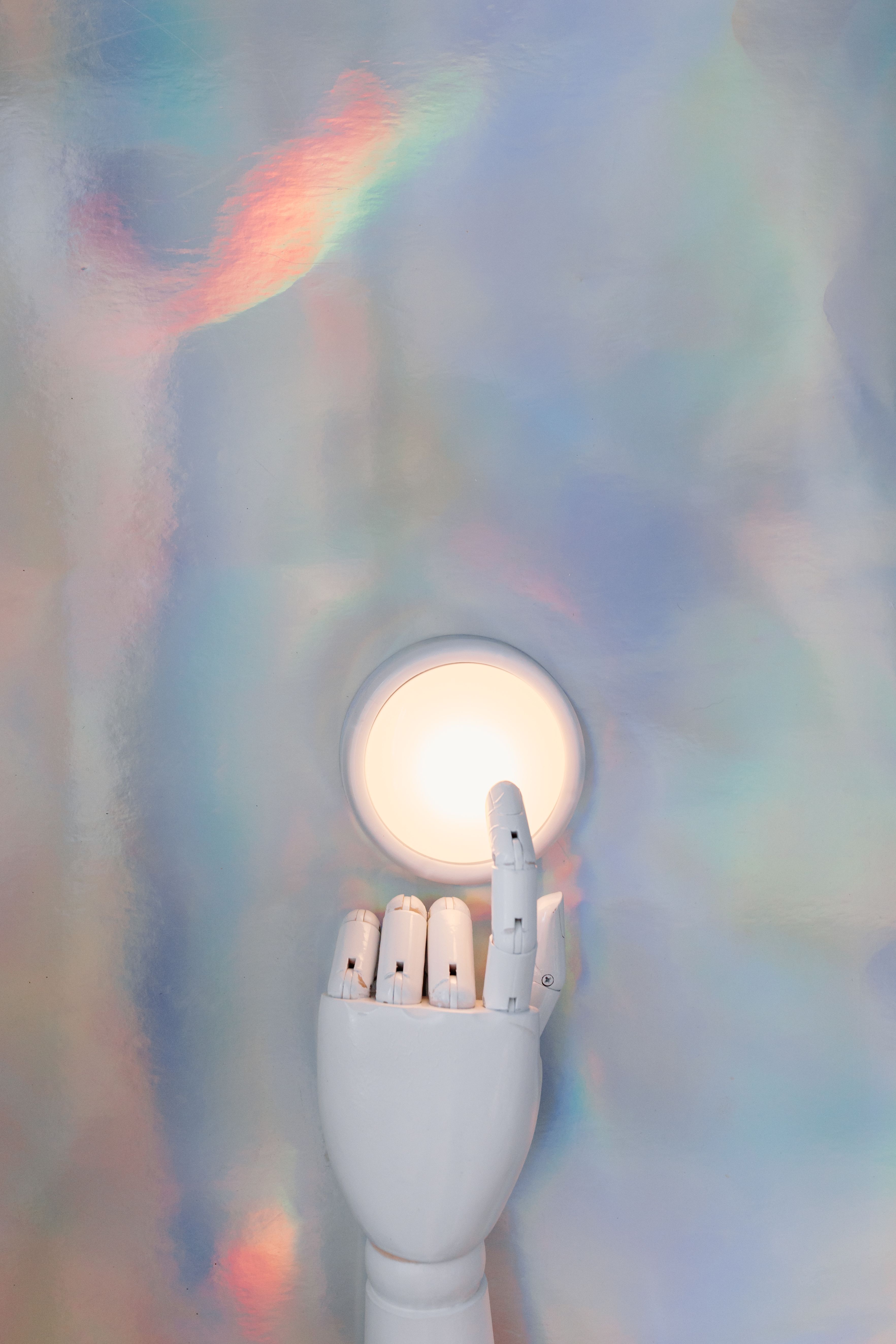 A pale articulated hand reaching up toward a small round glowing light on a soft iridescent background.
